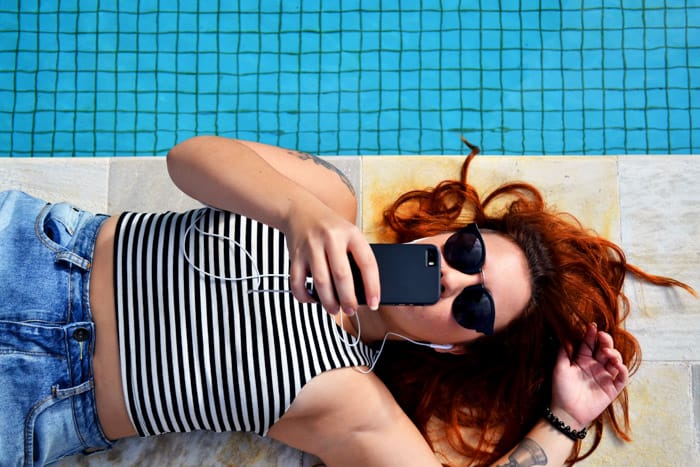 Overhead shot of a girl sunbathing by a swimming pool while editing photos on her smartphone 